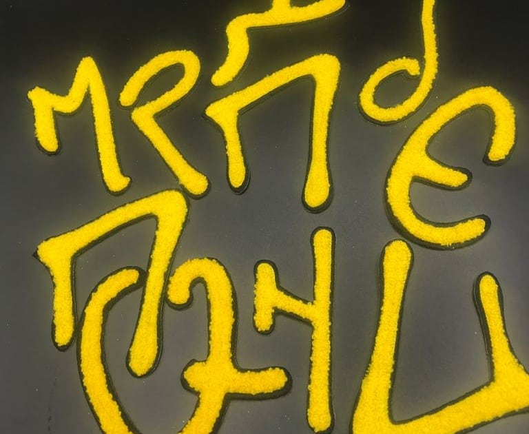 a black studio background shows multiple greek alphabets on the front as custom made chenille patches in yellow yarn with black embroidered trim outline