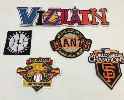 5 different custom made sports related embroidered patches on white background shows 11 inches wide big alphabet patch representing different America sports clubs in letters. The script overall says ‘VILLAIN’ with each letter shows off different league sport club like V anonymous, I stands for ‘Iowa Hawkeyes’, L stands for ‘Los Angeles Lakers’ over the traditional basketball in yellow, next L shows off for ‘Louisville Cardinals’, the capital A represents ‘Atlanta Angels’, another I reflects ‘Illinois Fighting Illini’ and the last letter big N linking to official logo and color representing ‘New York Mets’. The other four logos under it from the left number one is ‘San Francisco Giants Baseball Club’, number two in bottom square black twill over white thread embroider ‘LONGHORNS BASKETBALL’, the number three it’s left a logo for ‘Arter Member 1901,2001 American League 100 SEASONS, number four on deep right on the picture in custom made sport patch for 2010 World Series Champion.