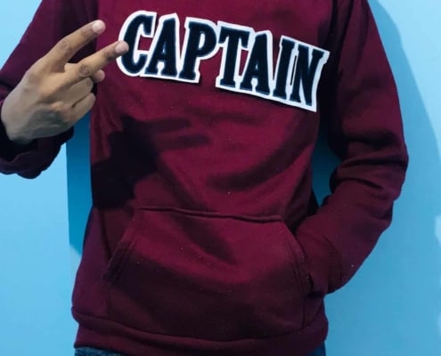 headless boy wearing maroon hoodie with white fabric black chenille patch on front saying CAPTAIN shows victory sign front of sky blue wall