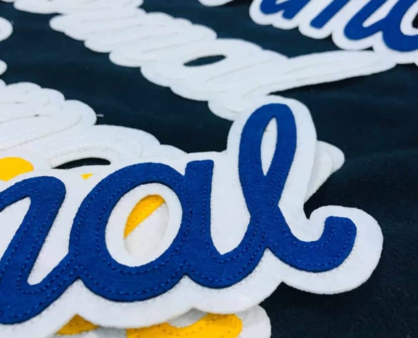 sets of custom made felt patches laser cut hot kiss in spread on black fabric on the back. Double felt patches show italic letters make script that say NORMAL. 1.White felt over white stitched in same color thread 2. Blue felt over white stitched in blue color thread 3. Yellow felt over white stitched in yellow color thread