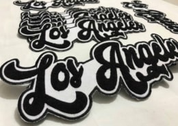 a set of five 10 inches wide chenille embroidered made custom script patches that say LOS ANGELES in black yarn on white scrim felt. The four patches lay down to the floor while the fifth shows off front of white background studio. Featuring black embroidered outline on white fabric and beautiful curve font fluffy city name of great California.