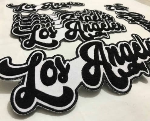 a set of five 10 inches wide chenille embroidered made custom script patches that say LOS ANGELES in black yarn on white scrim felt. The four patches lay down to the floor while the fifth shows off front of white background studio. Featuring black embroidered outline on white fabric and beautiful curve font fluffy city name of great California.