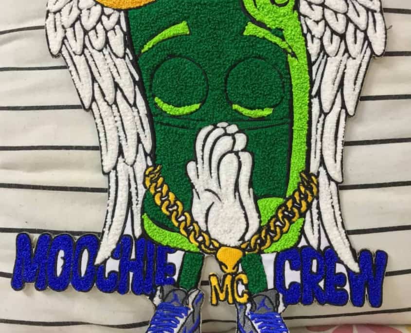 big size chenille art patch in two sheds of green chenille keeping hand in prayer; chains around and locket named MC in gold satin; white fluffy wings both sides and gold yellow ring on hand. Wearing stylish sneakers and text over ankles reads MOOCHIE on left and CREW on right in blue yarn over check background