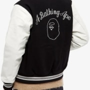 boy shows up back of letterman jacket in black with an artwork in white and text saying A BATHING APE , white sleeves