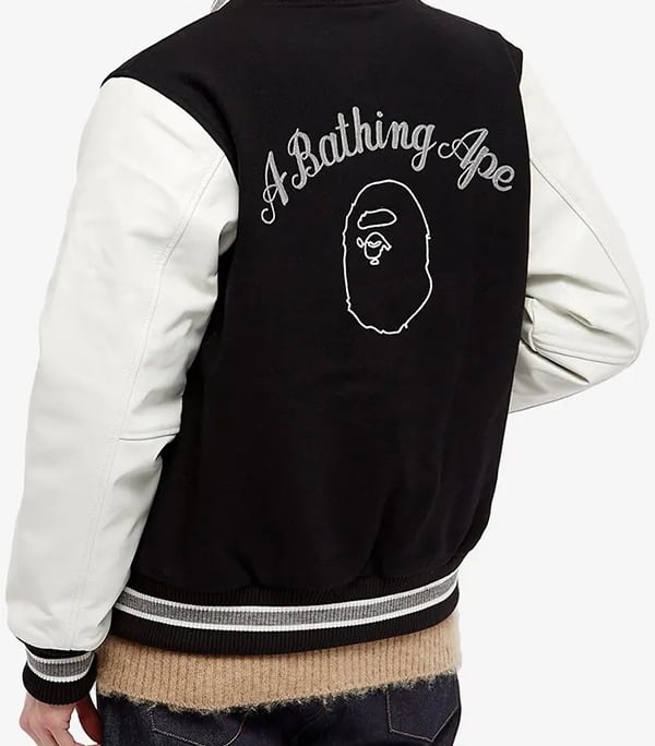 boy shows up back of letterman jacket in black with an artwork in white and text saying A BATHING APE , white sleeves