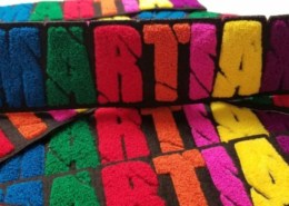 Bulk chenille felt patches in multi color spread that says ‘MARTIAN’ in caps lock capital English alphabet. M in blue yarn, A in green, R in red, T in orange, I in pink, A in yellow and N in fluffy purple on the black scrim felt and twill. The script letters show cut off scars passing through entire wording.