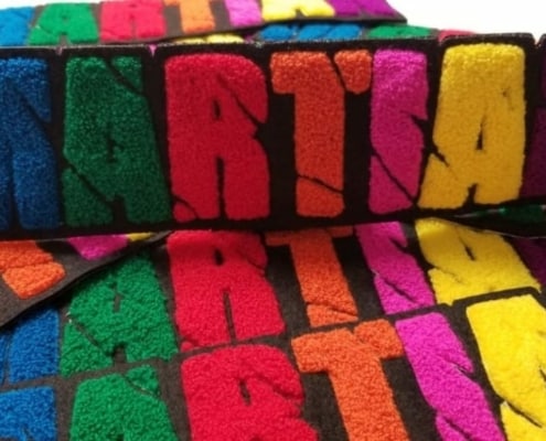 Bulk chenille felt patches in multi color spread that says ‘MARTIAN’ in caps lock capital English alphabet. M in blue yarn, A in green, R in red, T in orange, I in pink, A in yellow and N in fluffy purple on the black scrim felt and twill. The script letters show cut off scars passing through entire wording.