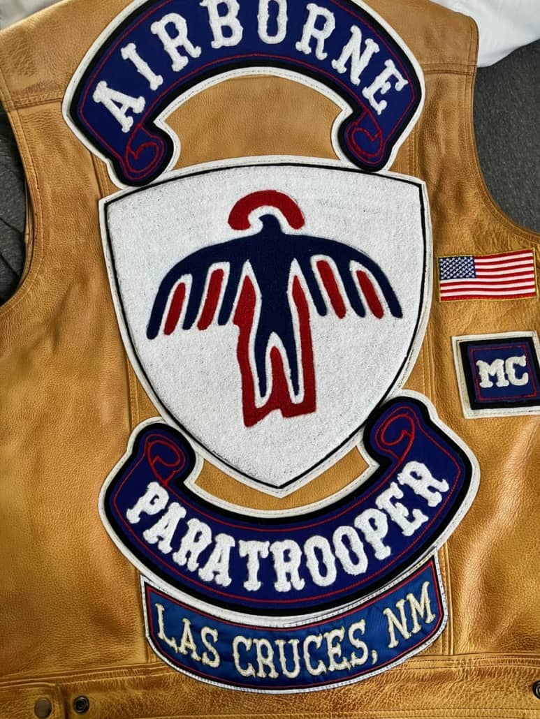 A set of big shield shape and rockers patches on light brown motorcyclists leather vest, a shield in the center shows navy blue and red design while white chenille filled around and a black thin line of embroidery, a big rocker on the top on double applique white/black fabric and white chenille letters says AIRBURNE, a same size and style rocker in bottom of shield says PARATROOPER, a small 4” square patch on the side of vest says MC in white chenille on double felt of white and black, another rocker in most bottom on silk fabric in blue says LAS CRUCES, NM