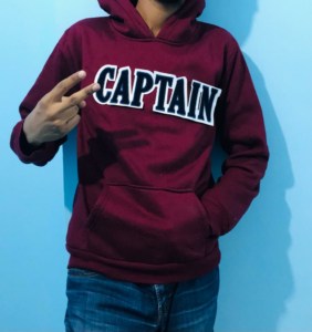 boy hiding head on blue background wall shows victory sign wearing maroon hoodie, blue jeans and white felt fluffy chenille text patch says CAPTAIN in navy blue