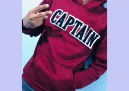 head less boy wearing maroon hoody show victory sign from left hand and right hand inside pocket showing chenille letter patch on the front on white fabric and navy chenille inside lettering that reads C A P T A IN