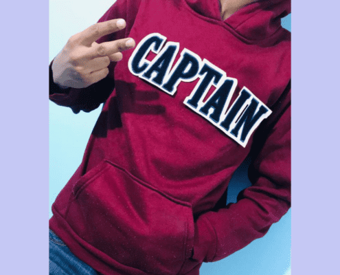 head less boy wearing maroon hoody show victory sign from left hand and right hand inside pocket showing chenille letter patch on the front on white fabric and navy chenille inside lettering that reads C A P T A IN