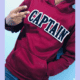 head less boy wearing maroon hoody show victory sign from left hand and right hand inside pocket showing chenille letter patch on the front on white fabric and navy chenille inside lettering that reads C A P T A IN